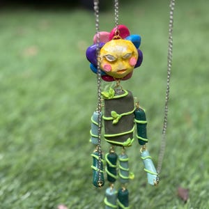 May include: A hanging mobile sculpture featuring a central figure with a yellow sun-shaped head surrounded by multi-colored petals. The body is composed of dark green cylindrical segments wrapped in light green vines, suspended by thin silver chains.