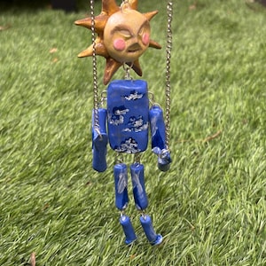 May include: A hanging mobile featuring a sculpted sun head with gold rays and pink cheeks, connected by silver chains to a blue rectangular torso and limbs decorated with white cloud patterns. The figure is suspended by three thin silver chains.
