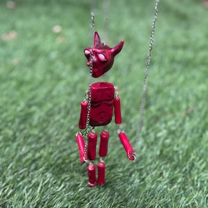 Puede incluir: Un collar con colgante articulado de color rojo que representa una figura con cuernos similar a un diablo, con la cabeza y el torso separados. Las extremidades están formadas por segmentos cilíndricos unidos por finas cadenas metálicas plateadas.
