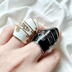 May include: Two statement rings are shown, one white and one black. The white ring features gold detailing and a decorative emblem. The black ring has silver accents and a similar emblem. Both rings have a distinctive, layered design.