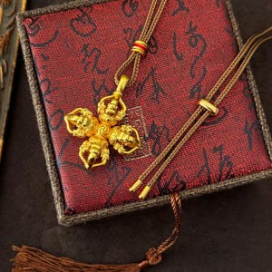 May include: A gold pendant necklace with a four-sided design, on a red patterned box. The necklace has a brown cord with gold accents and a tassel. The box has a dark red background with a black pattern.