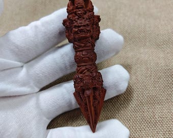 May include: A reddish-brown carved wooden phurba featuring intricate details of a wrathful deity face at the top, a central cylindrical section with decorative patterns, and a pointed blade base adorned with dragon-like motifs and animal carvings.