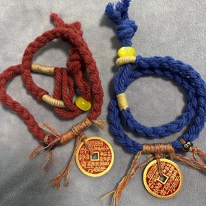 Puede incluir: Dos pulseras de cuerda trenzada, una roja y otra azul, cada una adornada con un colgante de moneda dorada. La pulsera roja presenta una cuenta amarilla y detalles dorados, mientras que la azul tiene una cuenta similar y detalles dorados. Ambas tienen borlas.
