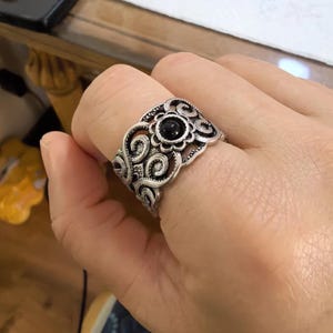Könnte beinhalten: Ein breiter Ring aus silberfarbenem Metall mit einem komplizierten Filigranmuster und geschwungenen Verzierungen. In der Mitte des Bandes befindet sich ein kleiner, runder, glatter schwarzer Stein in einer gebogten, blütenförmigen Fassung.