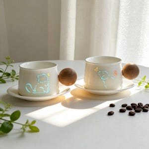 May include: Two white ceramic mugs with matching saucers, each featuring a round wooden handle. One mug displays a light blue cat outline with a butterfly, while the other shows a light blue dog outline with a butterfly. Coffee beans are scattered nearby.