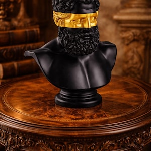 Könnte beinhalten: Schwarze Büste eines Mannes mit Bart und Locken, verziert mit einer goldenen Augenbinde. Die Skulptur steht auf einem dunklen Holztisch mit kunstvollen Schnitzereien. Dekorativer Artikel.