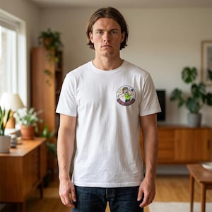 Peut inclure: Un t-shirt blanc à col rond avec un graphisme coloré sur le devant. Le graphisme représente un personnage de dessin animé jouant au golf avec le texte "Bender Peterson" dans un design circulaire. Le t-shirt est porté par une personne dans une pièce avec des meubles en bois.