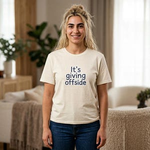 Peut inclure: Un t-shirt à manches courtes de couleur crème avec une texture mouchetée et le texte bleu foncé "It's giving offside" imprimé au centre de la poitrine. Le t-shirt présente une encolure ronde et une coupe standard, porté avec un jean bleu foncé.