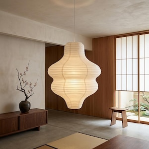Peut inclure: Une grande lanterne en papier blanc cassé est suspendue au plafond dans une pièce aux accents de bois. La lanterne a une forme unique et superposée et émet une lueur douce et chaleureuse. La pièce présente un design minimaliste avec de la lumière naturelle.