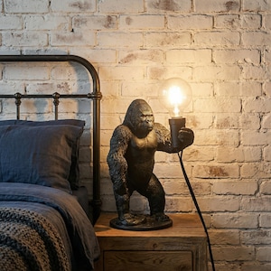 Puede incluir: Una lámpara de mesa negra con forma de gorila que sostiene una bombilla redonda. La lámpara está sobre una mesita de noche de madera junto a una cama con una colcha y almohada azul oscuro. El fondo es una pared de ladrillo blanco.