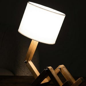 May include: A wooden desk lamp with a white lampshade. The lamp's base and support structure are made of light-colored wood, with joints allowing for adjustable positioning. The lampshade is cylindrical and emits a soft glow, creating a warm ambiance.