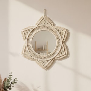 May include: A star-shaped, beige macrame mirror hangs on a cream-colored wall. The mirror has a circular center and a fringed edge. A wooden table below holds a vase with dried flowers, folded towels, and a potted succulent.