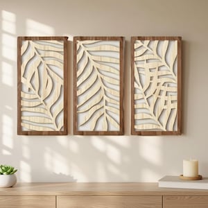 Peut inclure: Ensemble de trois panneaux muraux en bois. Chaque panneau présente un motif de feuilles de couleur claire dans un cadre marron foncé. Les panneaux sont affichés sur un mur clair, au-dessus d'un meuble en bois, avec une plante succulente et une bougie.