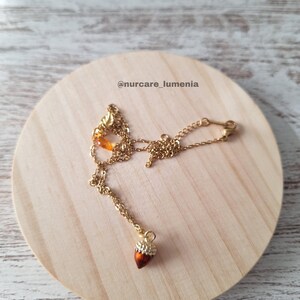 K&ouml;nnte beinhalten: Eine goldfarbene Halskette mit einem Eichelanh&auml;nger. Der Eichelanh&auml;nger ist braun mit einer goldfarbenen Kappe und mit kleinen, klaren Kristallen verziert. Die Halskette wird auf einer runden, hellen Holzoberfl&auml;che pr&auml;sentiert. Der Text "@nurcare_lumenia" ist sichtbar.