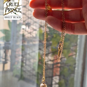 K&ouml;nnte beinhalten: Eine goldfarbene Halskette mit einem Eichelanh&auml;nger. Die Kette wird vor einem Fenster gehalten, im Hintergrund ist das Buch "The Cruel Prince" von Holly Black zu sehen.