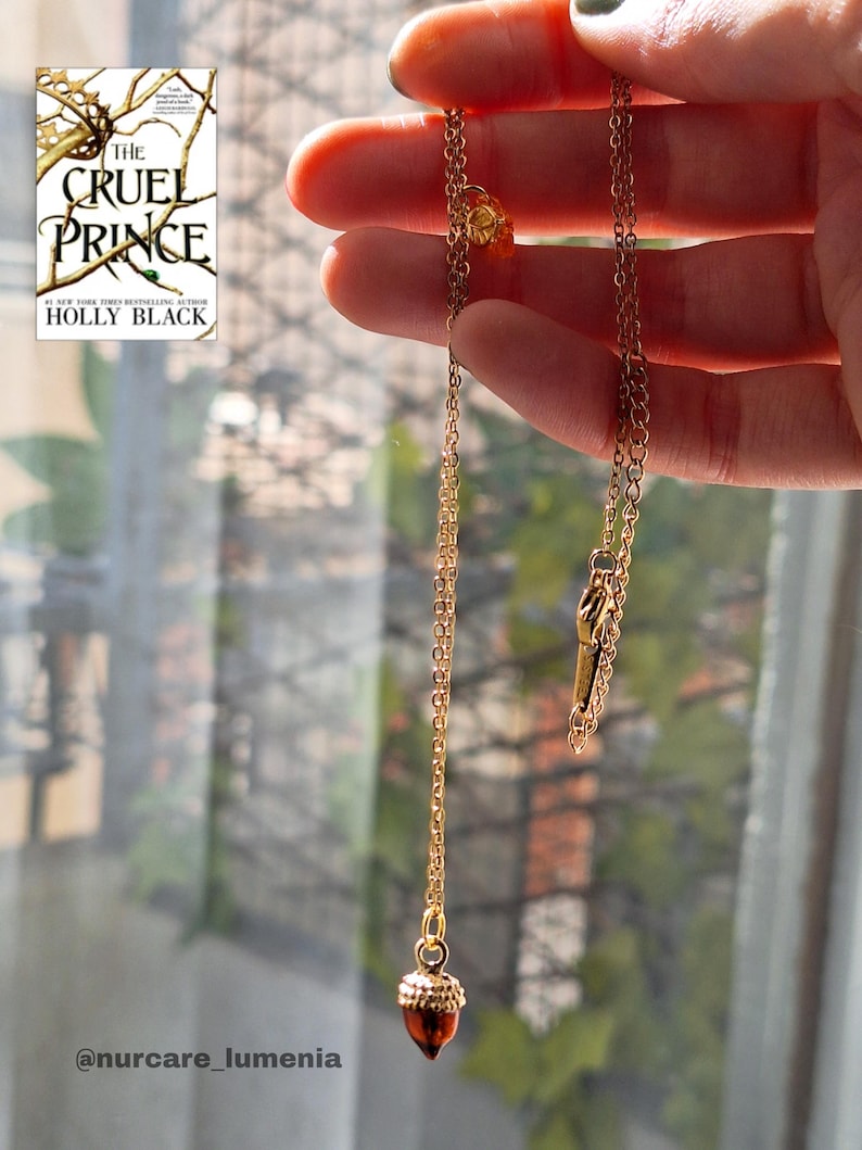 K&ouml;nnte beinhalten: Eine goldfarbene Halskette mit einem Eichelanh&auml;nger. Die Kette wird vor einem Fenster gehalten, im Hintergrund ist das Buch "The Cruel Prince" von Holly Black zu sehen.