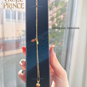 K&ouml;nnte beinhalten: Eine goldfarbene Halskette mit einem kleinen, orangefarbenen Anh&auml;nger in einer schwarzen Schachtel. Die Kette hat eine feine Kette und einen Blatt-Charm. Auf der Schachtel steht der Text "THE CRUEL PRINCE" in Goldbuchstaben.
