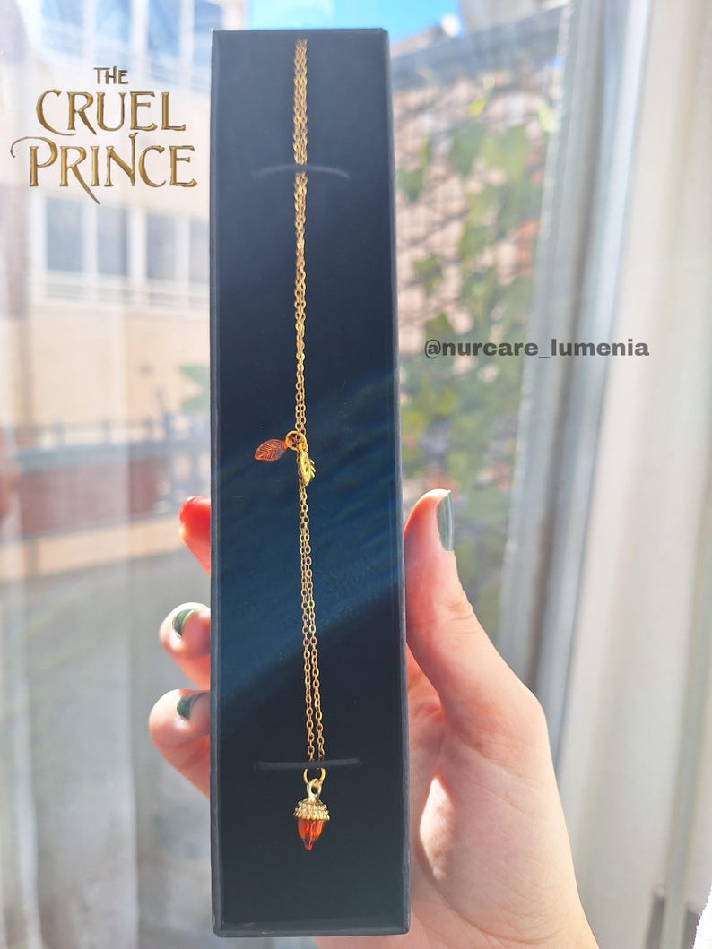 K&ouml;nnte beinhalten: Eine goldfarbene Halskette mit einem kleinen, orangefarbenen Anh&auml;nger in einer schwarzen Schachtel. Die Kette hat eine feine Kette und einen Blatt-Charm. Auf der Schachtel steht der Text "THE CRUEL PRINCE" in Goldbuchstaben.