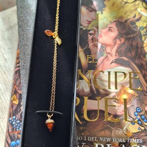 K&ouml;nnte beinhalten: Ein goldfarbener Lesezeichen mit Kette und Eichelanh&auml;nger, pr&auml;sentiert in einer schwarzen Box. Das Lesezeichen hat Blatt- und Eichel-Charms. Das Buchcover zeigt eine romantische Illustration und den Titel "El Principe Cruel".