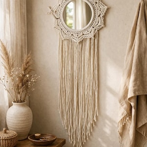 Può includere: Uno specchio rotondo con una cornice in macramè color crema e lunghe frange è appeso a una parete beige. Un vaso con fiori secchi, una ciotola di legno e un cestino di vimini sono su un tavolo di legno. Un asciugamano beige è appeso alla parete.