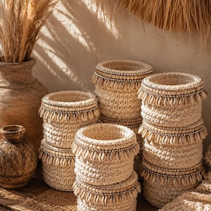 Peut inclure: Plusieurs paniers crochetés de couleur crème, ornés de coquillages et de perles. Les paniers cylindriques de différentes hauteurs sont disposés sur une surface en bois. Des plantes séchées et des poteries sont en arrière-plan.