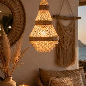 Op de afbeelding: Een beige hanglamp met een conische vorm, versierd met geweven details en een gloeilamp aan de binnenkant. Het licht hangt boven een tafel met een kaars en een vaas. Een geweven spiegel en een macramé wandkleed zijn ook zichtbaar.