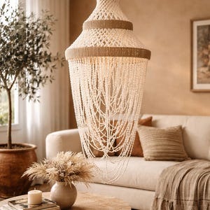 May include: A cream-colored, tiered chandelier with a woven jute trim and beaded fringe hangs above a round wooden coffee table. The room features a neutral color palette with a sofa, throw pillows, and a potted olive tree.
