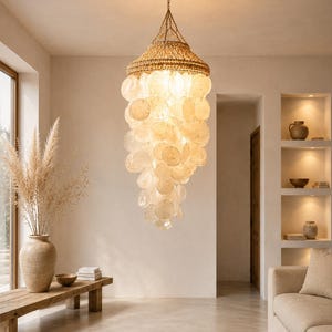 Peut inclure: Un lustre à étages composé de nombreux disques circulaires translucides en nacre capiz, suspendus à une structure en fibres tressées de couleur naturelle. Le luminaire diffuse une lueur chaude et douce, avec des coquillages en cascade.