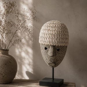 May include: A decorative sculpture of a human head featuring a textured, speckled gray surface and a cap covered in numerous small, white, oval-shaped cowrie shells. The head is mounted on a thin black metal rod attached to a square black base.