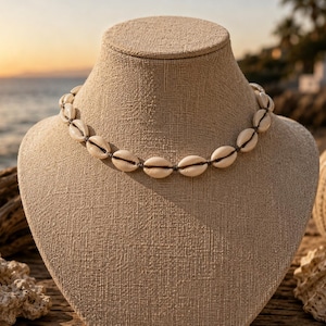 Peut inclure: Un collier de coquillages crème, présenté sur un buste de mannequin beige. Le collier est composé de coquillages ovales reliés par un cordon foncé. L'arrière-plan comprend un coucher de soleil sur la mer, du bois flotté et des poteries décoratives.