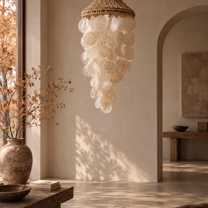 Op de afbeelding: Een gelaagde kroonluchter met talloze doorschijnende, ronde capiz-schelpschijven die aan een geweven, natuurkleurig vezelframe hangen. De lamp hangt aan een dunne ketting en heeft een trapsgewijs toelopend silhouet van gelaagde schelpen.