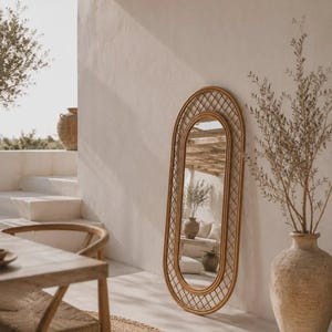 Puede incluir: Un espejo ovalado de suelo con un marco de ratán marrón claro que presenta un patrón de celosía decorativo alrededor del cristal central. El espejo está colocado en posición vertical contra una pared, reflejando el espacio y la luz.