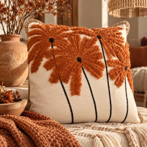 May include: A square throw pillow with an off-white base featuring three embroidered palm tree designs in a textured, burnt orange thread. The palm trunks are represented by thin, vertical black lines, and the pillow has a fringed edge.