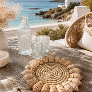 Peut inclure: Un dessous de verre rond en matériau tressé, orné de coquillages. Une bouteille d'eau en verre transparent et deux verres assortis sont posés sur une table en bois. En arrière-plan, une plage et la mer.
