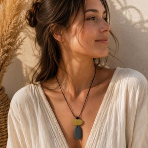 Peut inclure: Un collier avec un cordon noir et deux formes de pendentif : un petit ovale jaune moutarde au-dessus d'un ovale gris plus grand. Le collier est porté sur un haut crème. L'arrière-plan est beige neutre.