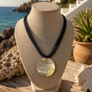 Op de afbeelding: Een ketting met een ronde, iriserende schelpenhanger aan een zwart koord. De ketting wordt getoond op een beige buste. De achtergrond toont een kustlandschap met een blauwe zee en lucht.