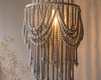 Op de afbeelding: Een gelaagde kroonluchter met een wit metalen frame versierd met talloze strengen lichtgrijze houten kralen. De kralen zijn gerangschikt in gedrapeerde, geschulpte patronen die verticaal hangen, wat een gelaagd effect creëert rond een centrale lichtbron.