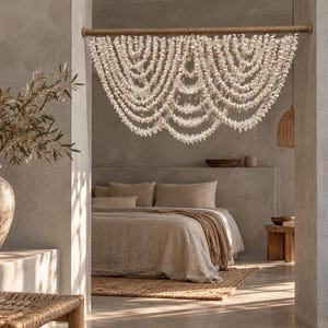 Puede incluir: Un tapiz de pared compuesto por múltiples hileras drapeadas de pequeñas conchas blancas suspendidas de una barra de madera horizontal. Las conchas están dispuestas en una serie de niveles festoneados superpuestos, creando un efecto de cortina.