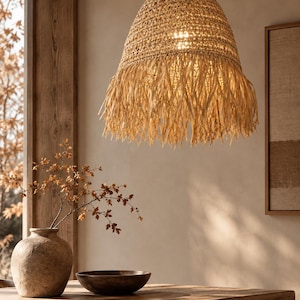 May include: A bell-shaped pendant light fixture crafted from woven natural tan fibers, featuring a textured upper dome and a long, loose fringe trim at the base. The light hangs from a thin black cord, emitting a warm glow from within the weave.