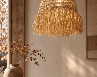 Op de afbeelding: Een klokvormige hanglamp gemaakt van geweven natuurlijke vezels in een beige kleur, met een getextureerde bovenkoepel en een lange, losse franje aan de onderkant. De lamp hangt aan een dun zwart snoer en verspreidt een warme gloed door het vlechtwerk.