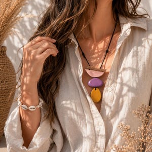 Peut inclure: Un collier avec un cordon noir présente trois pendentifs en bois colorés : un demi-cercle rose, un ovale violet et une goutte jaune. Un bracelet en coquillages blancs est également visible. La personne porte une chemise beige boutonnée.