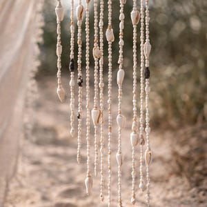 Op de afbeelding: Een decoratief schelpengordijn hangt tegen een wazige achtergrond van zand en groen. Het gordijn is gemaakt van strengen kleine, ronde kralen afgewisseld met schelpen van verschillende maten en vormen. De schelpen zijn voornamelijk crèmekleurig met enkele donkere accenten.