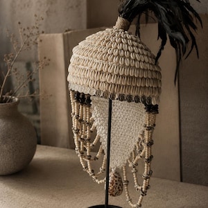 Peut inclure: Une coiffe décorative ornée de rangées de coquillages cauris crème, dotée d'un panache de plumes noires au sommet, d'une bande de petites pièces argentées et de brins pendants de perles en bois clair avec un coquillage tacheté unique.