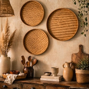 Peut inclure: Trois plateaux ronds en rotin tressé sont accrochés à un mur beige texturé. Une table en bois présente des ustensiles de cuisine, dont un vase en céramique, des cuillères en bois et une planche à découper. Un abat-jour tissé est suspendu au plafond et une plante est suspendue au mur.