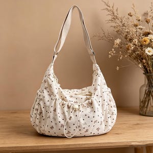 May include: Cream-colored shoulder bag with a repeating pattern of small brown hearts and teddy bears. The bag has a drawstring closure, two side pockets, and a long, adjustable strap. The bag is made of a soft, corduroy-like material.