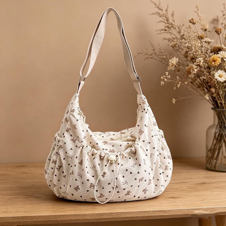 May include: Cream-colored shoulder bag with a repeating pattern of small brown hearts and teddy bears. The bag has a drawstring closure, two side pockets, and a long, adjustable strap. The bag is made of a soft, corduroy-like material.