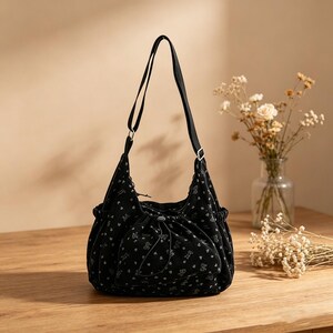 May include: Black shoulder bag with a drawstring closure and a long, adjustable strap. The bag features a repeating pattern of small, light-colored floral and heart shapes. The bag is sitting on a wooden surface.