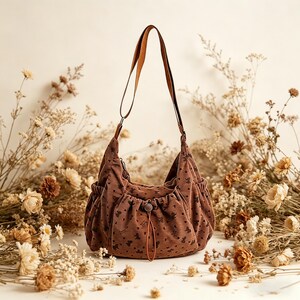 May include: A brown corduroy shoulder bag with a floral pattern and a brown leather strap. The bag has a drawstring closure and two side pockets. The bag is surrounded by dried flowers.