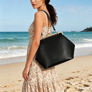 May include: A black handbag with a unique geometric shape and a gold-colored clasp. The bag has a long black strap and is being carried by a person wearing a floral dress on a beach.