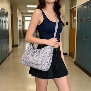 May include: A light gray shoulder bag with a drawstring closure and a pattern of small hearts and butterflies. The bag has a matching gray strap and is being carried over the shoulder.